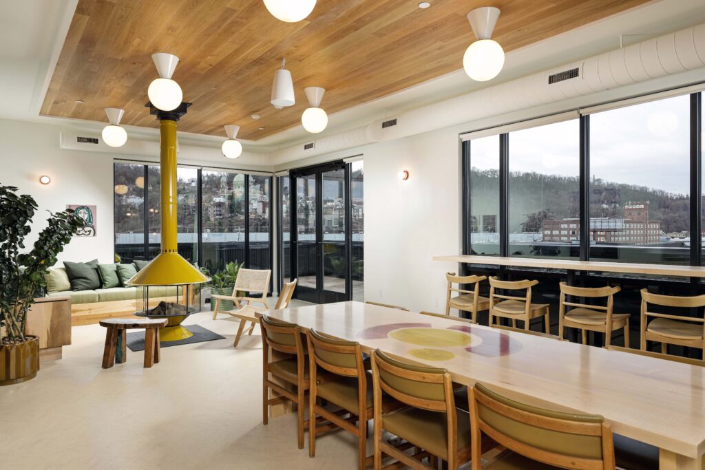 A modern lounge area with a yellow conical fireplace, a long wooden dining table, and large windows overlooking a city skyline.