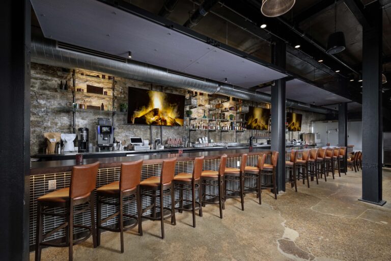 A long bar with rows of brown leather stools, exposed stone walls, and flat-screen televisions showing a fireplace.