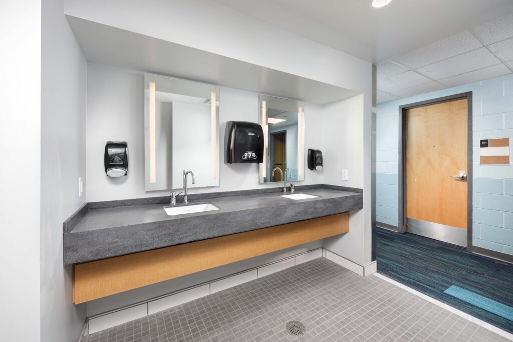 A public restroom vanity with two sinks, illuminated mirrors, and paper towel dispensers, with a wooden door visible in the background.