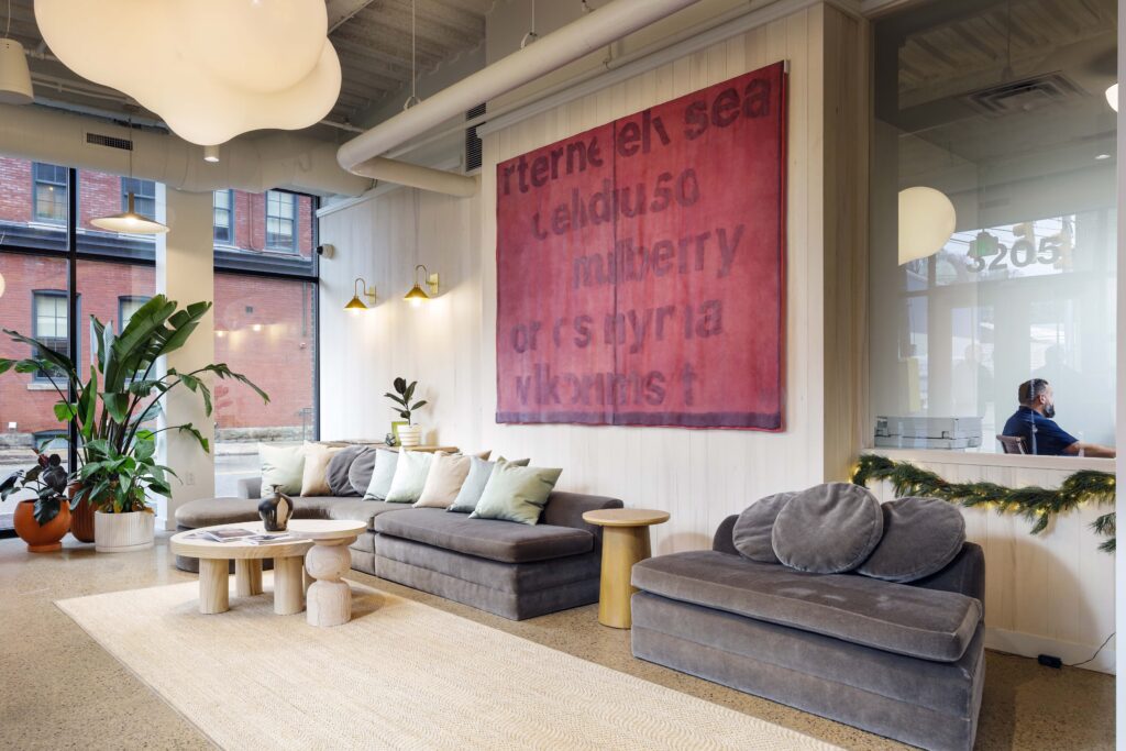 A modern lounge area with a long gray sofa, a separate chaise lounge, wooden coffee tables, and a large red wall hanging with black text.