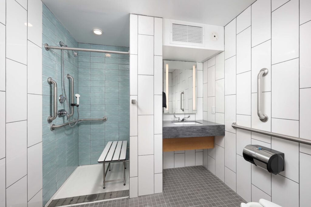 Accessible bathroom with a walk-in shower containing a bench and grab bars, next to a sink with an integrated mirror.