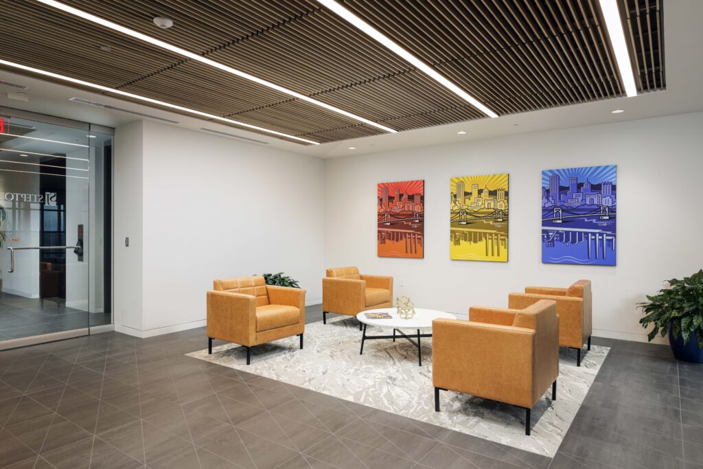 A lobby seating area with four tan leather armchairs arranged around a round white coffee table on a patterned rug, beneath three colorful city skyline artworks.