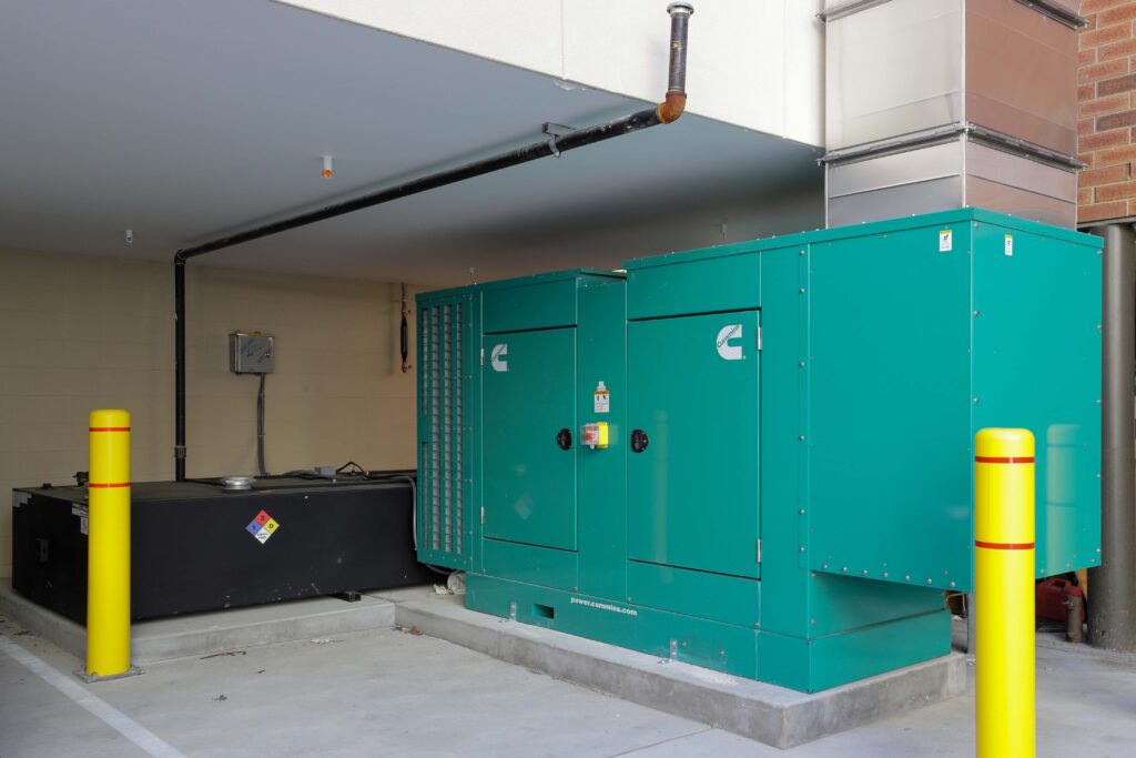 A large green Cummins industrial generator sits next to a black fuel tank in a concrete outdoor enclosure with yellow protective bollards.