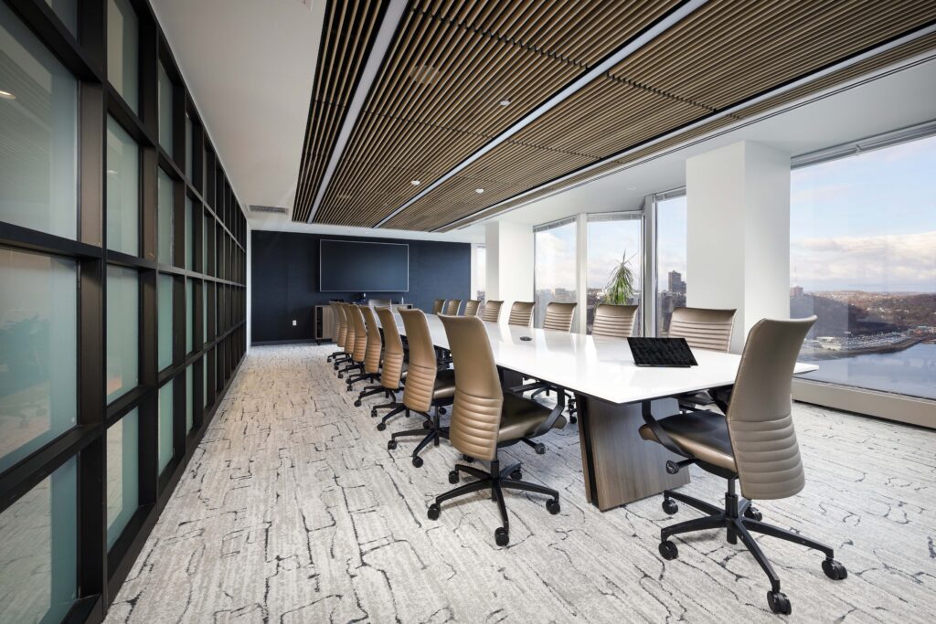 A modern boardroom with a long white table, twelve tan chairs, and floor-to-ceiling windows overlooking a city skyline.