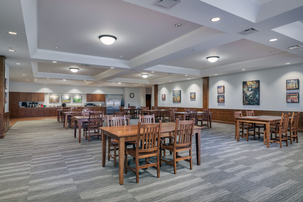 Dining hall with rows of wooden tables and chairs, a kitchenette in the background, and framed artwork on the walls.