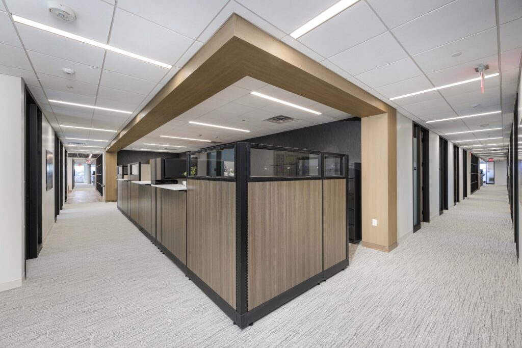 A row of office cubicles with wood-paneled partitions in a brightly lit office corridor.