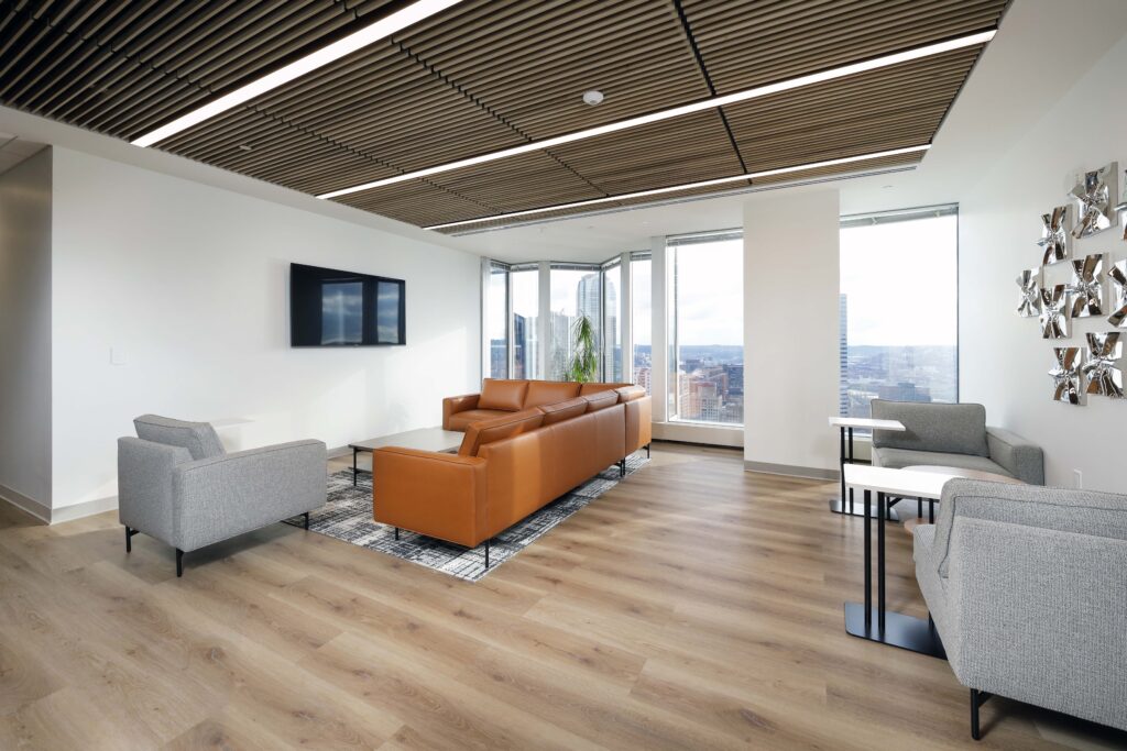 Modern office lounge with a tan leather sofa, grey armchairs, a television mounted on the wall, and floor-to-ceiling windows overlooking a city.