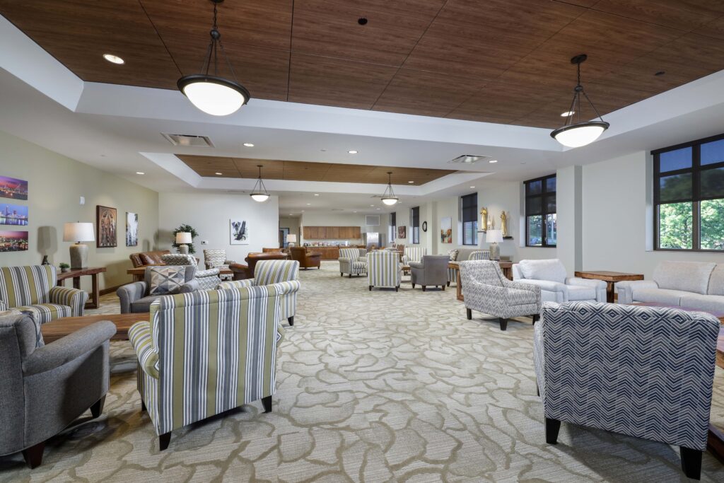 A large, open lobby featuring multiple seating areas with upholstered armchairs arranged on patterned carpeting, with pendant lighting and wood-paneled ceilings.