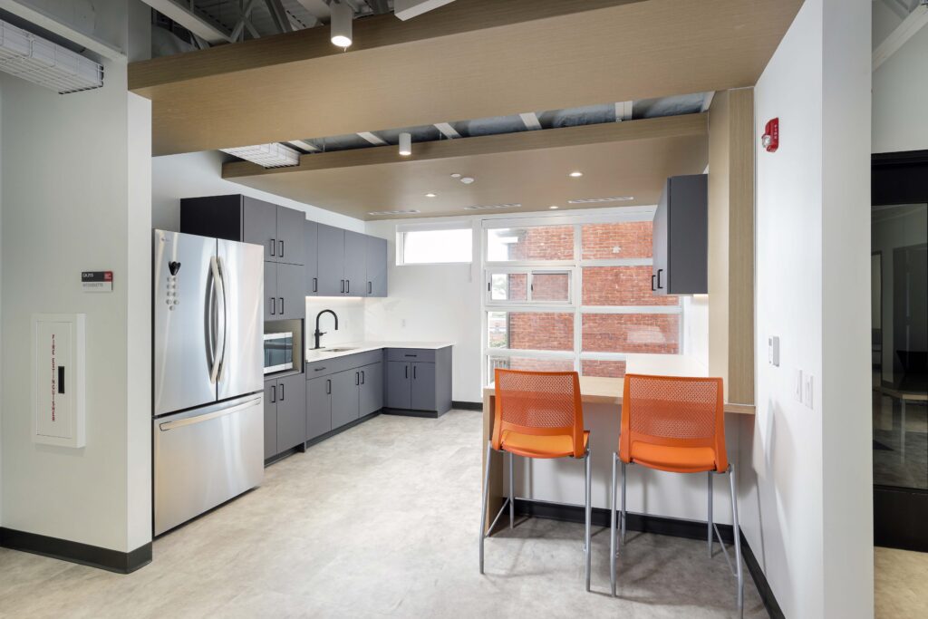A modern office kitchenette with dark grey cabinetry, a stainless steel refrigerator, and two orange stools at a breakfast bar.