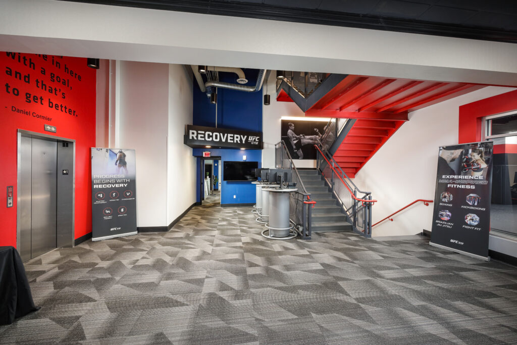 The entrance to a UFC Gym featuring a red staircase, a “Recovery” signage area, and two informational banners listing fitness programs and services.