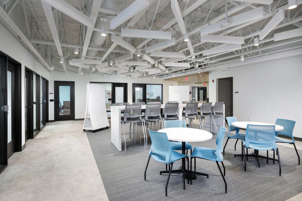 Modern office breakroom with a high communal counter, blue chairs at round tables, and an exposed ceiling with white acoustic baffles.