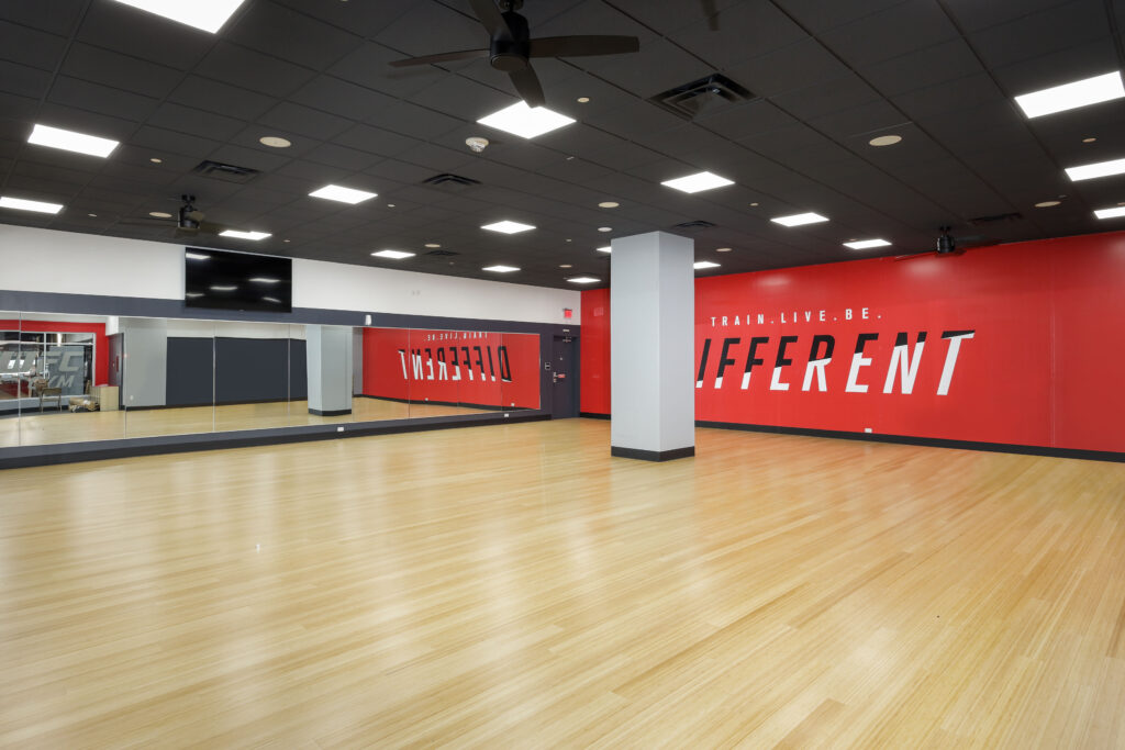 An empty gym studio with light wood flooring, a mirrored wall, and a large red wall featuring the text “TRAIN. LIVE. BE. DIFFERENT.”