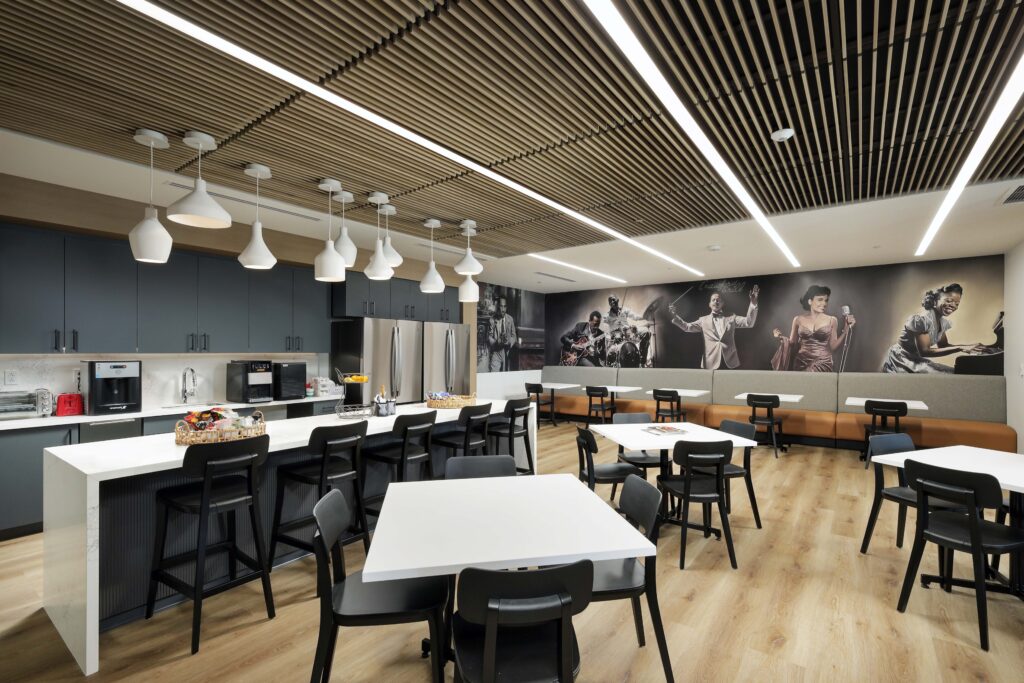 Modern office breakroom featuring a kitchen counter with bar stools, square dining tables with chairs, and a large mural of musicians on the back wall.