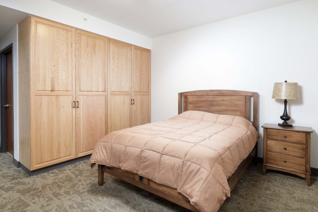 A bedroom with a wooden bed frame, a tan quilted comforter, a matching nightstand with a lamp, and floor-to-ceiling wooden storage cabinets.