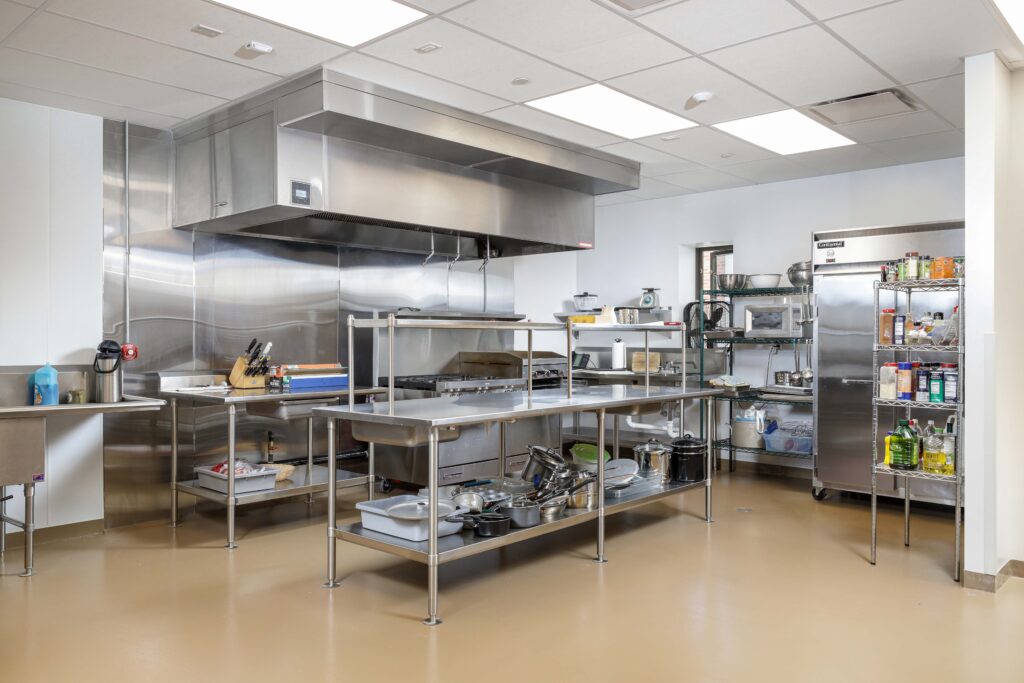 Commercial kitchen with stainless steel prep tables, a large exhaust hood over a range, and a metal shelving unit stocked with supplies.