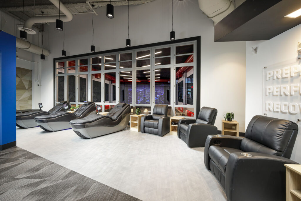 A wellness or recovery room featuring four reclining massage chairs and two black leather armchairs, with a sign on the wall reading “PREP PERF RECOV.”