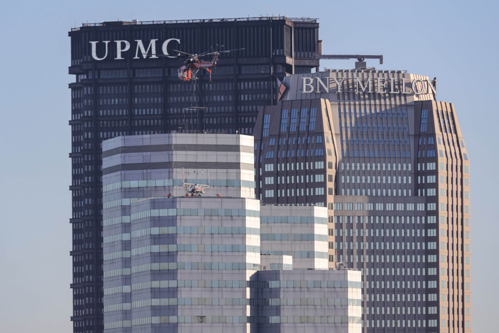 A helicopter uses a cable to lower equipment onto the roof of a skyscraper in Pittsburgh, with the UPMC and BNY Mellon buildings in the background.