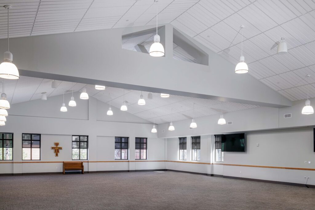 A large, empty hall with grey carpet, white walls, and multiple hanging pendant lights, featuring a wooden bench and a wall-mounted cross.