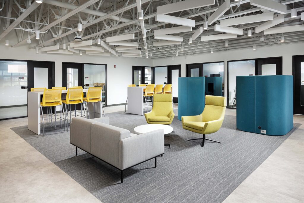 A modern office space featuring a gray sofa, two yellow armchairs, tall tables with yellow stools, and blue acoustic privacy partitions under an exposed ceiling.
