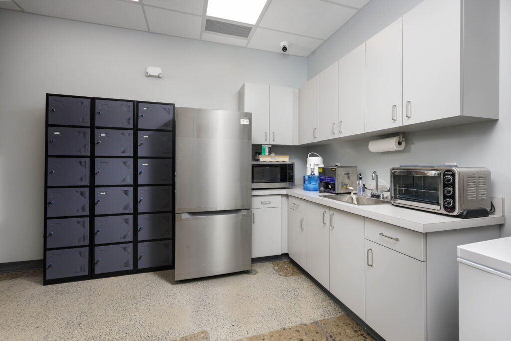 A breakroom featuring a bank of metal lockers, a stainless steel refrigerator, white cabinets, and a kitchenette counter with a sink and small appliances.