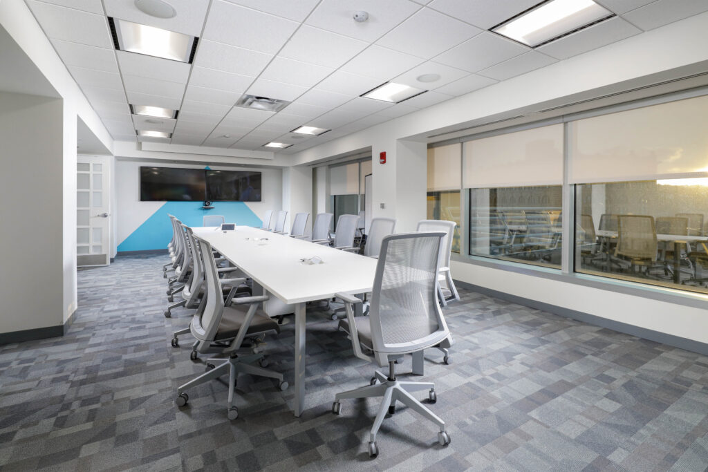 A modern conference room with a long white table, grey mesh chairs, and a wall-mounted flat-screen television.