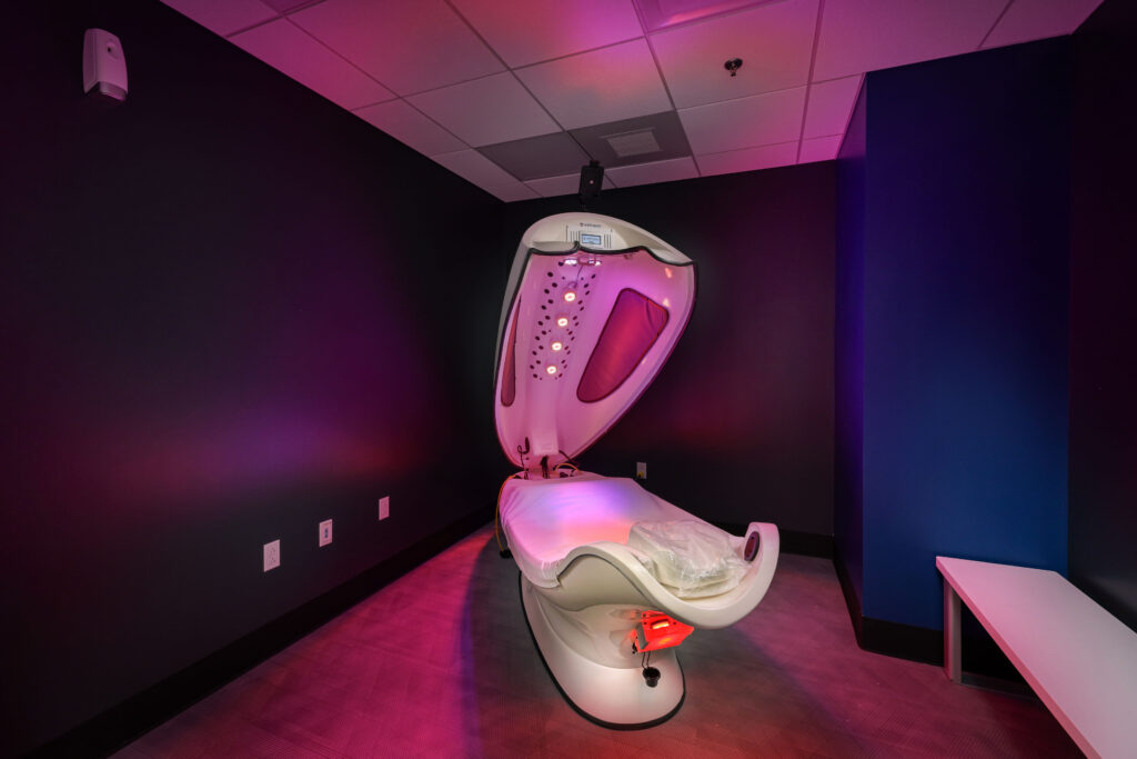 A wellness therapy pod with an open lid and interior lights, situated in a dark room with purple lighting.