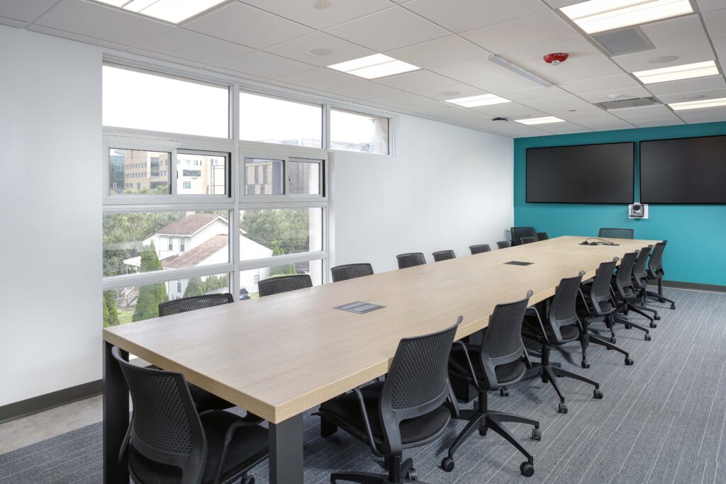 A long wooden conference table with black mesh office chairs in a modern meeting room with large windows and a teal accent wall.