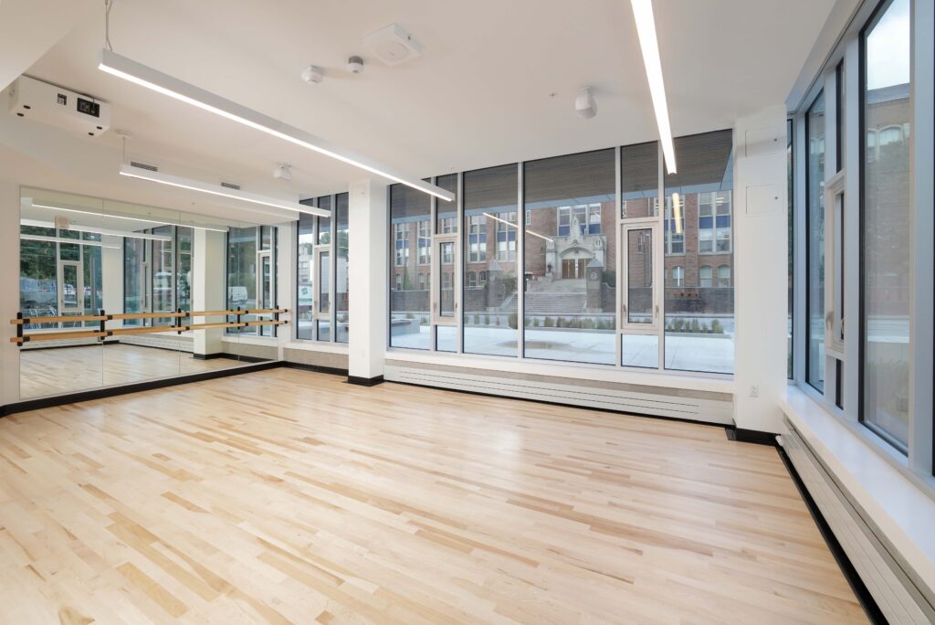 Empty dance studio with light wood floors, a wall of mirrors, and large windows overlooking a brick building.