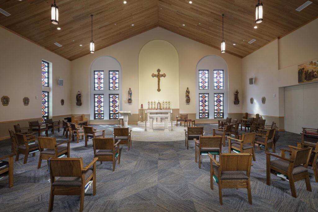 The interior of a church with rows of wooden chairs facing an altar, a crucifix, and four stained-glass windows.
