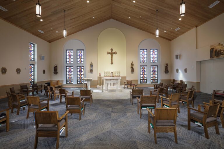 The interior of a church with rows of wooden chairs facing an altar, a crucifix, and four stained-glass windows.