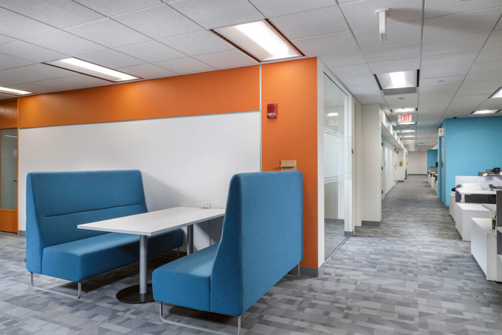 Modern office nook with two blue upholstered benches and a white table, positioned along an orange wall in a hallway.