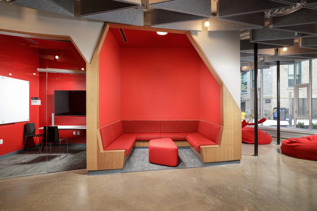 A recessed seating area with a red U-shaped booth and matching ottoman, next to a glass-walled study room with a white board and television.