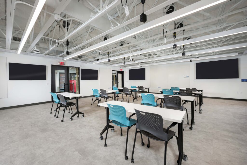 Modern classroom with rows of white desks and a mix of teal and black chairs, facing several wall-mounted television screens.