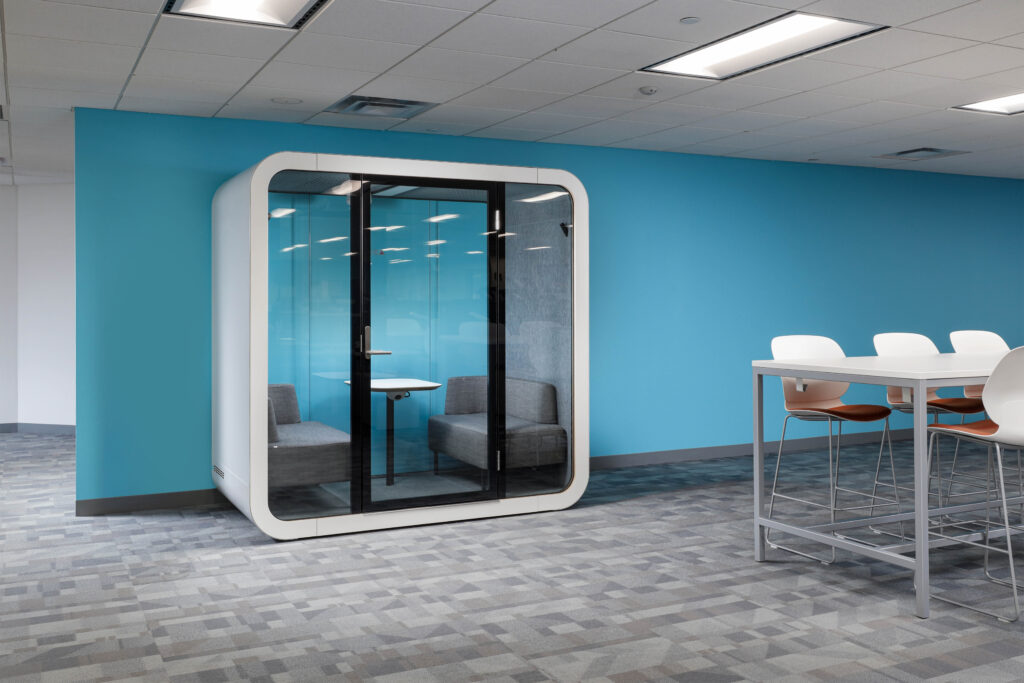A freestanding glass-walled meeting pod with two gray sofas and a small table, situated in an office against a blue wall next to a high-top table.