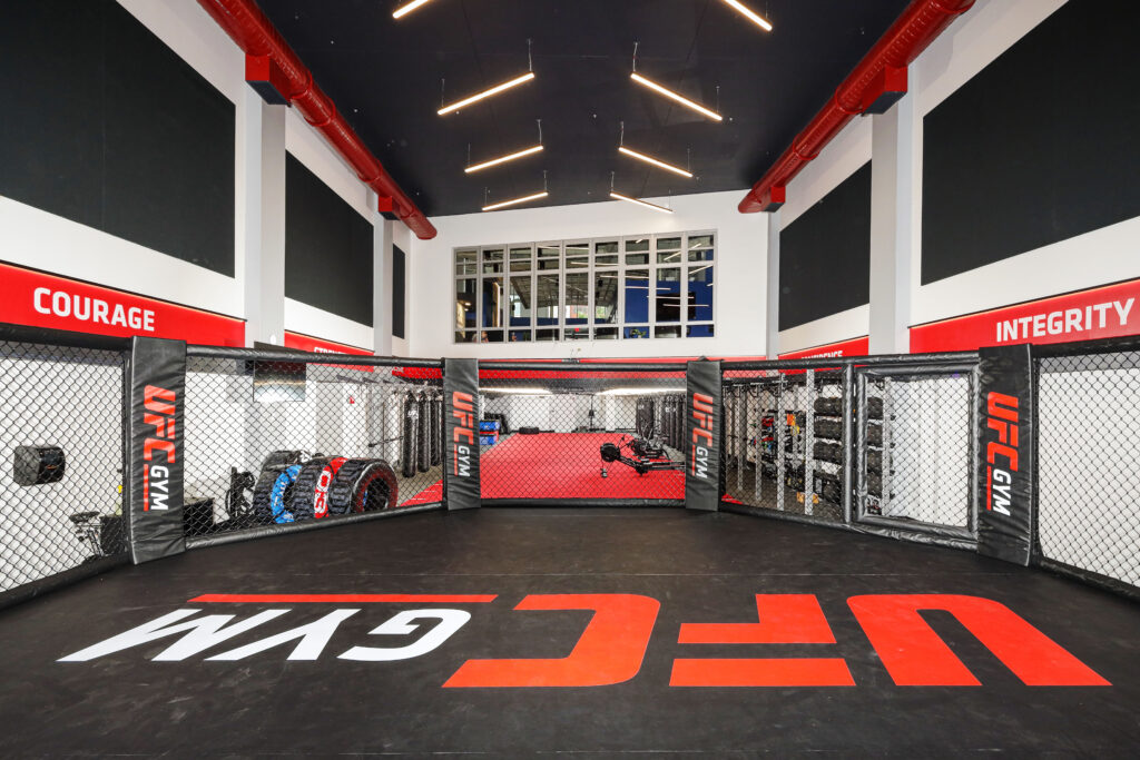 An octagonal mixed martial arts training cage with UFC Gym branding on the floor and padding.