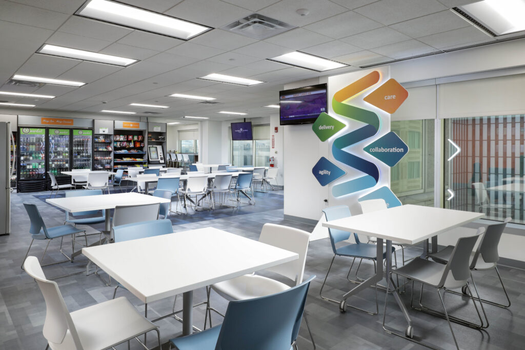 An office breakroom with white tables and chairs, a snack kiosk, and a wall display featuring a colorful graphic with the words “delivery,” “care,” “collaboration,” and “agility.”