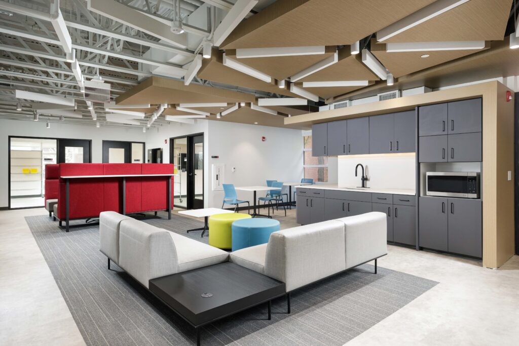 A modern office lounge featuring two grey sofas, a small kitchenette with grey cabinets, colorful circular ottomans, and a red privacy partition.