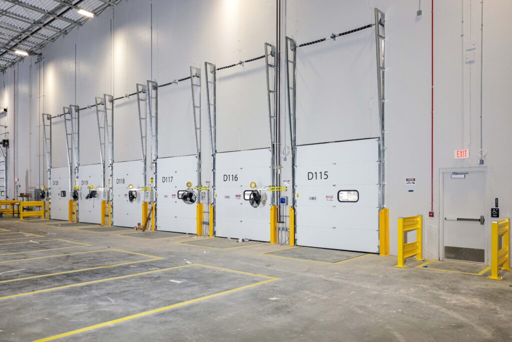 A row of numbered warehouse loading dock doors with yellow safety bollards and a single exit door on the far right.