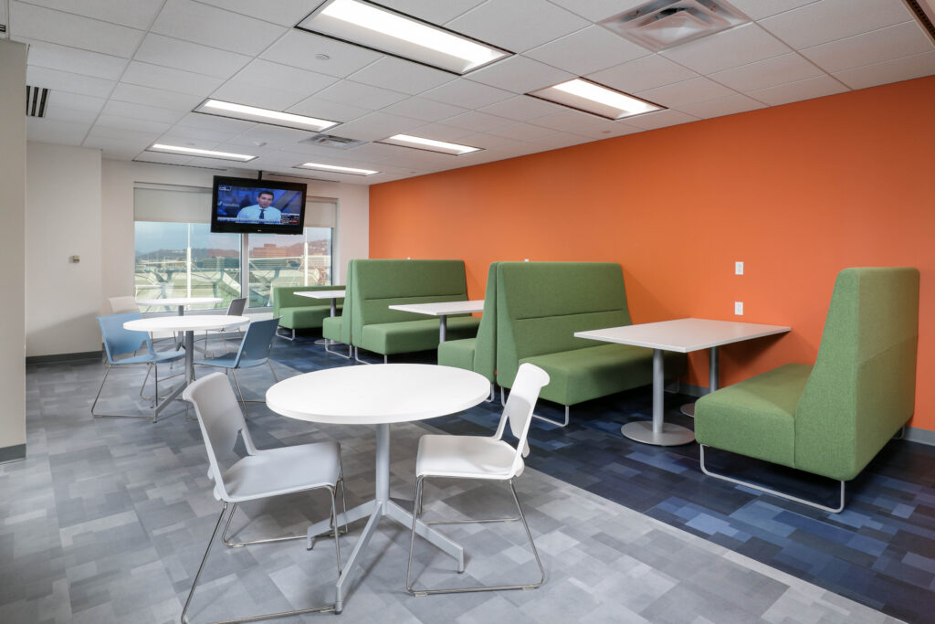 An office lounge area with green booths, white tables, and chairs, featuring a television mounted on the wall.