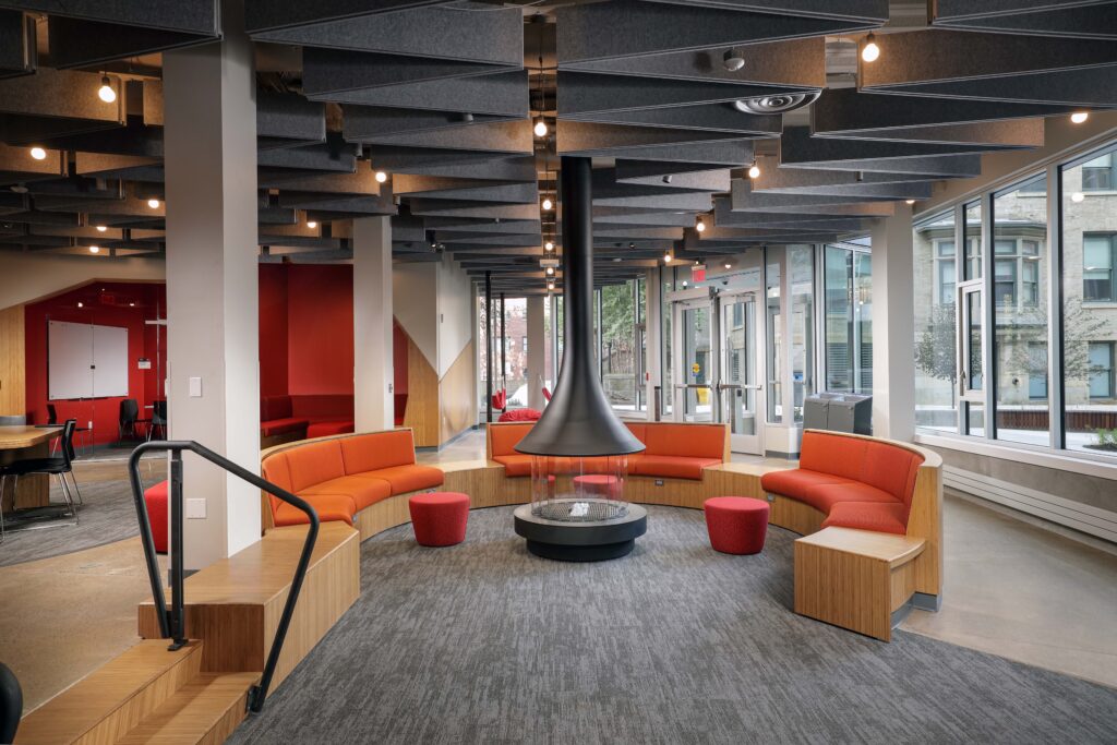 Curved orange seating arranged around a central suspended fireplace in a modern lounge area with geometric ceiling panels.