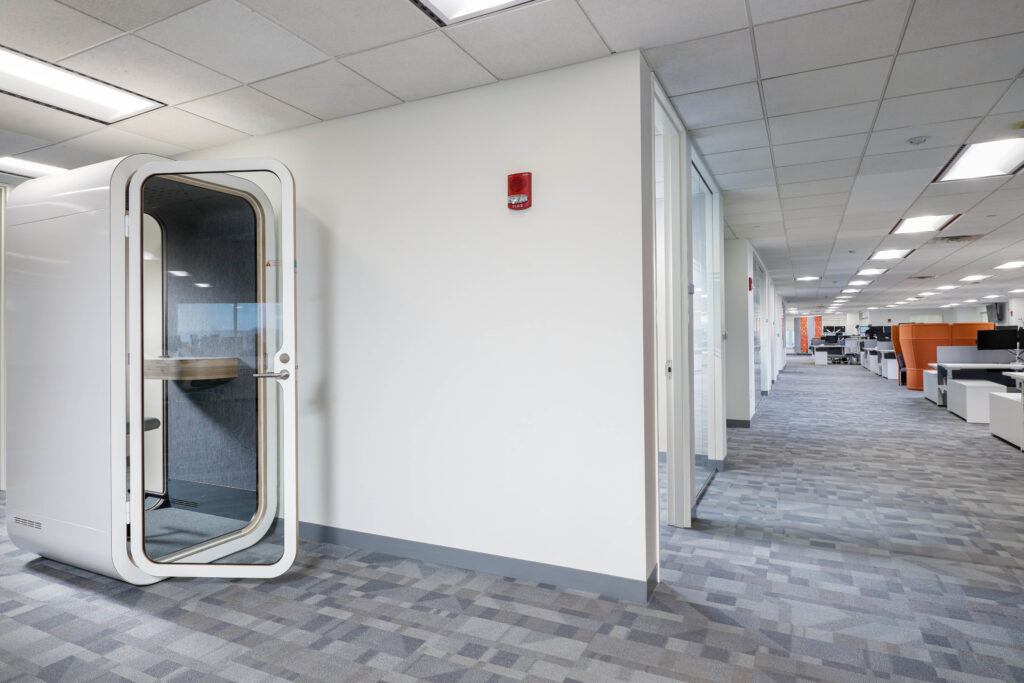 A white, rounded office phone booth situated in an open-plan office corridor.