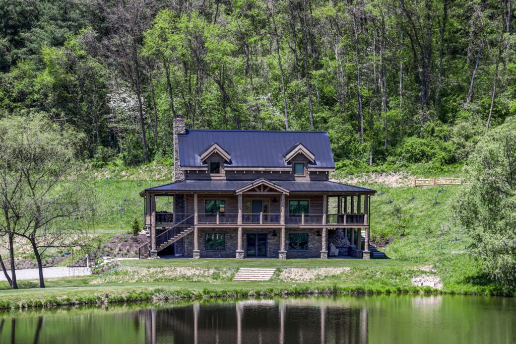 Two-story log cabin with a dark metal roof and wraparound porch, situated on a grassy hill overlooking a pond with a forest backdrop.