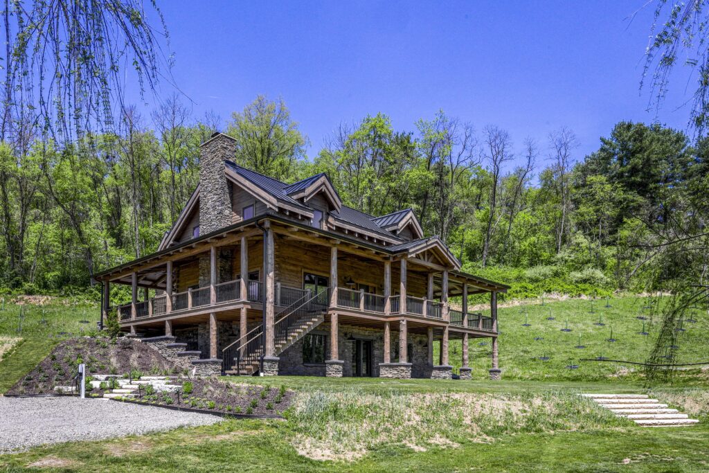 A two-story log cabin with a stone chimney and wraparound deck, situated on a grassy hillside backed by trees.