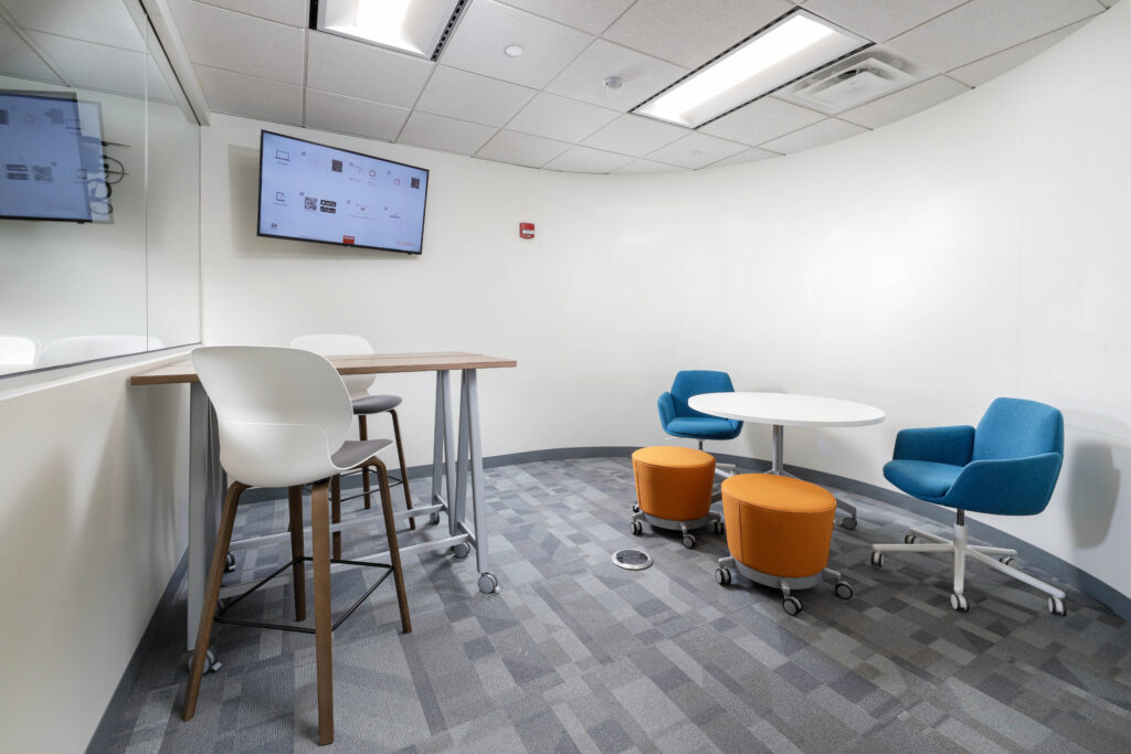 A small meeting room with a tall standing desk, two blue chairs at a round table, and two orange ottomans on wheels. A screen mounted on the wall displays a digital interface.