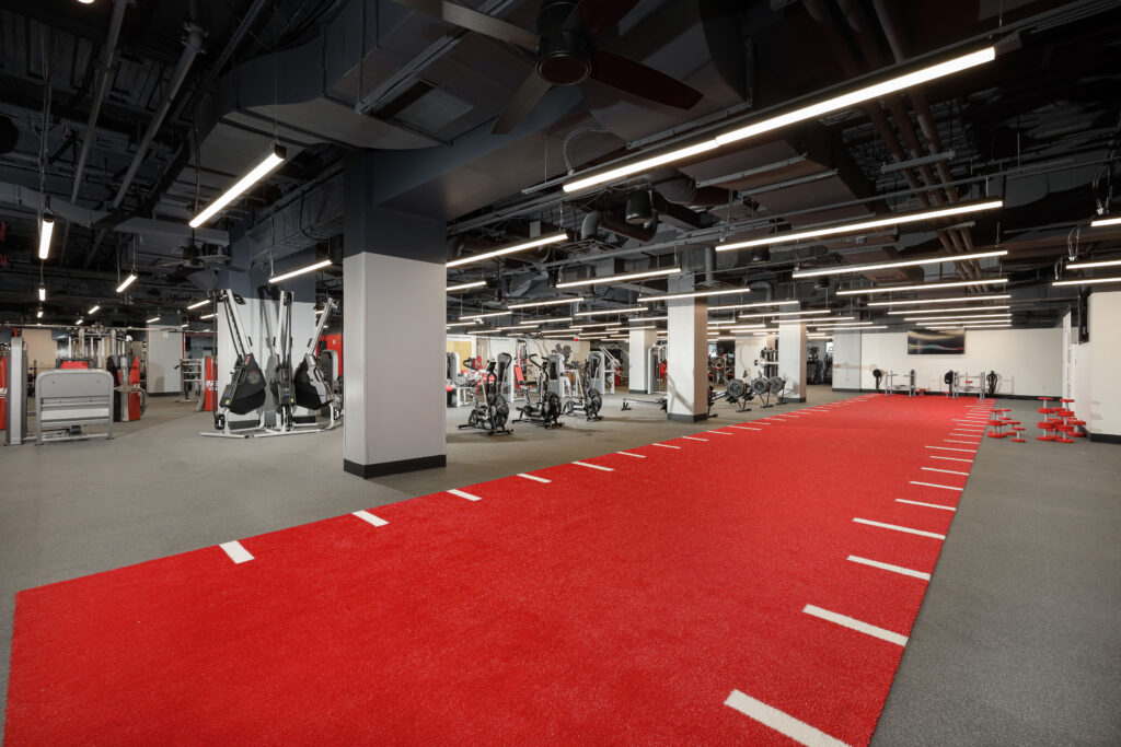 A gym floor featuring a long red turf track with white markings, surrounded by various fitness equipment.