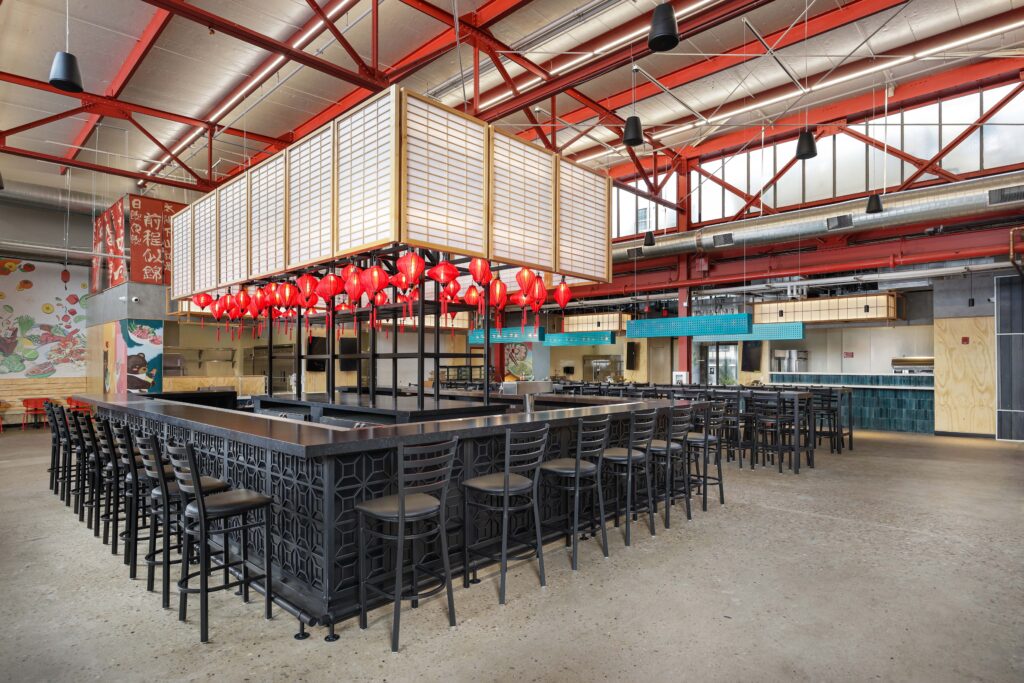 A restaurant interior featuring a central bar with black stools, illuminated by a grid of hanging red lanterns under a high, red-trussed ceiling.