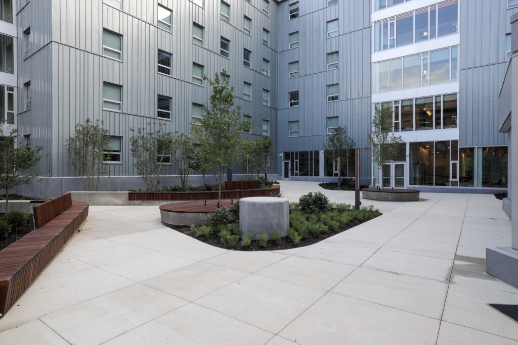 A modern building courtyard with light gray metal panels, concrete paths, wooden benches, and small trees.