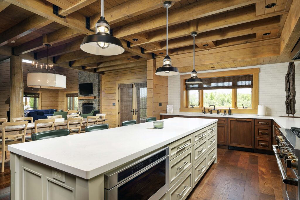 A kitchen with a large white island, wooden cabinets, and exposed ceiling beams, looking into a living room with a stone fireplace.