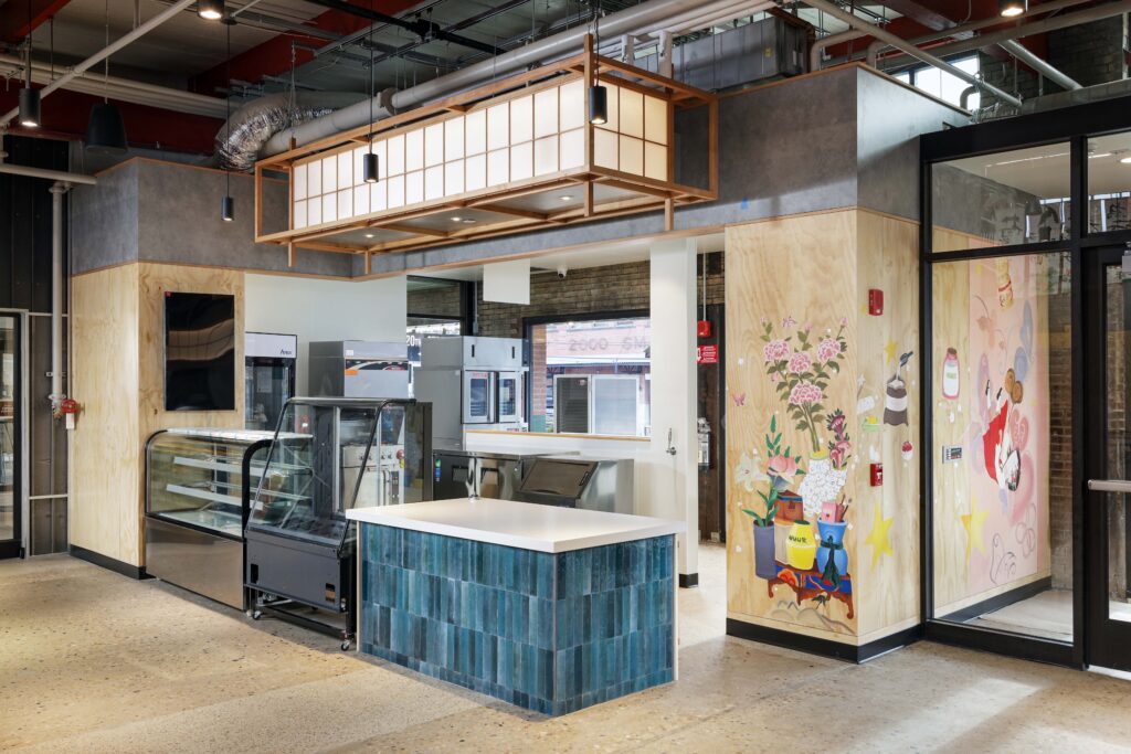 Interior of a cafe featuring a counter with blue tiles, glass display cases, and a wall mural of flowers and whimsical characters.