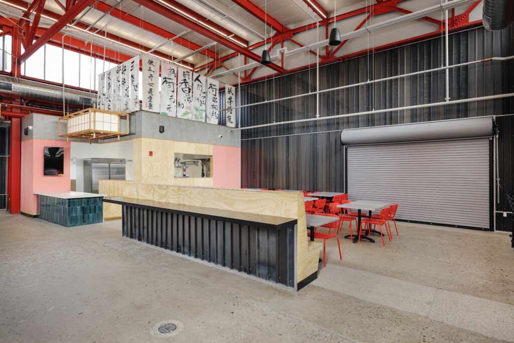 Interior of a restaurant with a plywood counter, red metal chairs, tables, and vertical banners with Asian script hanging from the ceiling.
